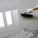 A gloved hand using a trowel to apply self-leveling compound to a concrete floor.