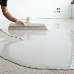 A person using a trowel to spread self-leveling compound on a concrete floor.