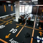A high-angle view of a gym floor covered in black rubber tiles with bright orange directional arrows, lines, and large white numbers indicating workout zones