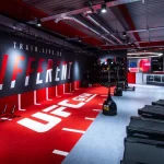 The floor of a UFC GYM featuring a prominent red area with the large white UFC GYM logo and grey flooring where treadmills are located.