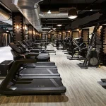 The floor of a well-equipped gym featuring a combination of light grey flooring in the foreground and wood-look planks in the background where cardio machines are placed.