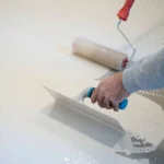 A close-up of a person's hand using a flat trowel to spread a thick, white, self-leveling compound or epoxy onto a floor, with a paint roller resting nearby on the wet surface.