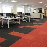 An open-plan office features a striking geometric carpet pattern in red, orange, and black, with numerous workstations.